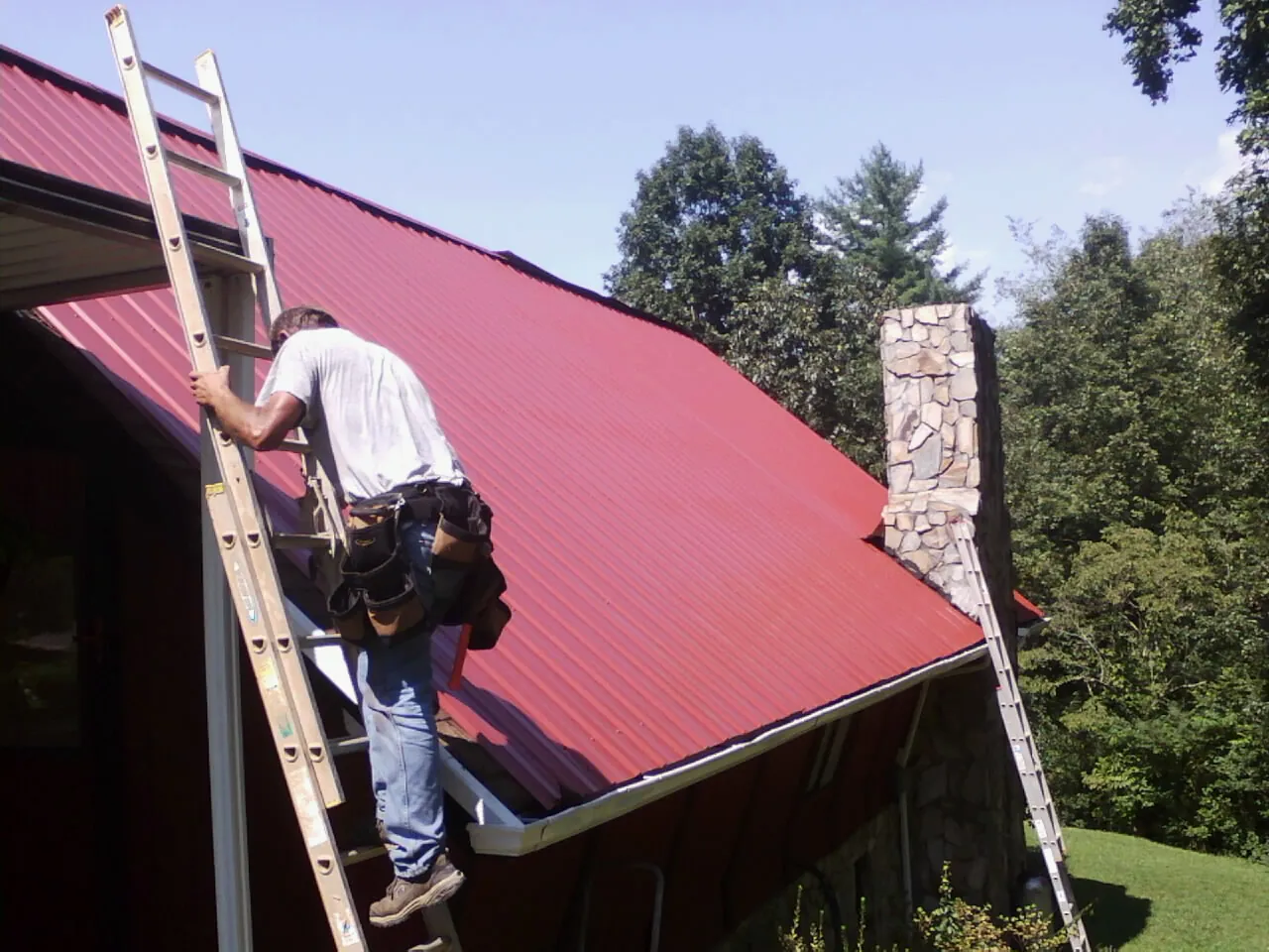 Roofer on a ladder beside a red metal roof and stone chimney — Soffit & Fascia Repair by Roofing Brothers in Jonesboro